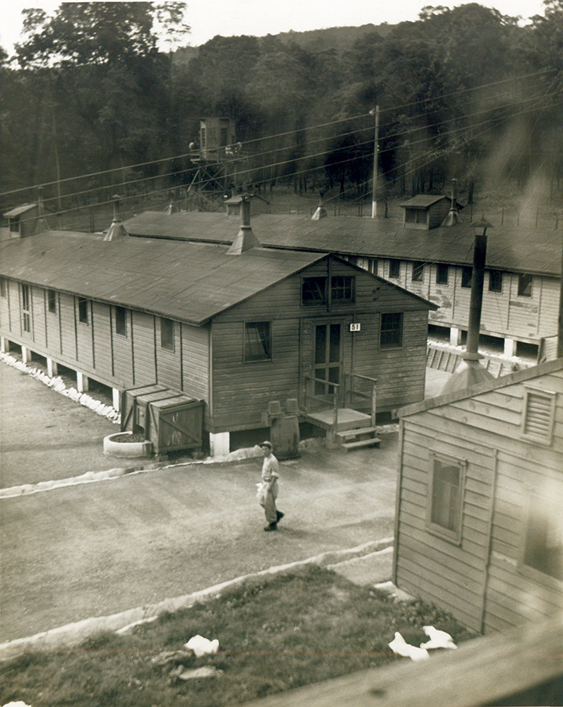 A prisoner walks through the barracks in Compound One at Pine Grove Furnace POW Interrogation Camp. A guard tower is visible in the background. Adams County Historical Society