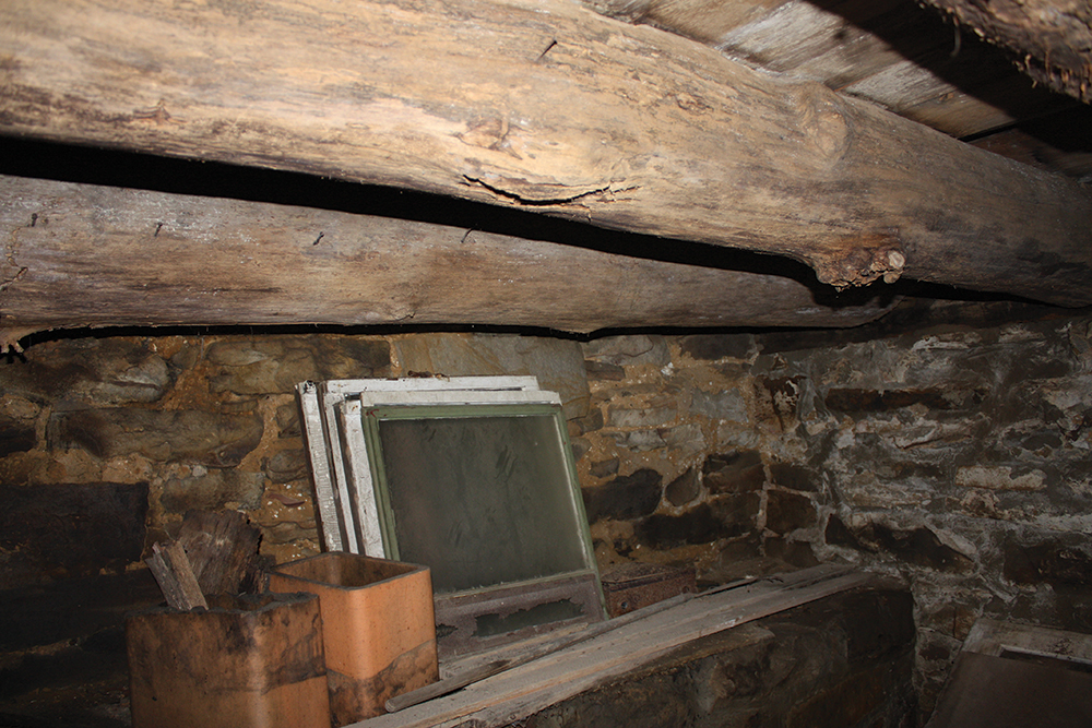 The log home’s cellar reveals construction at the north foundation wall, with hand-hewn log joists holding the floor above. The stones on the wall are larger at left to support the fireplace above. Photo, Katherine Peresolak