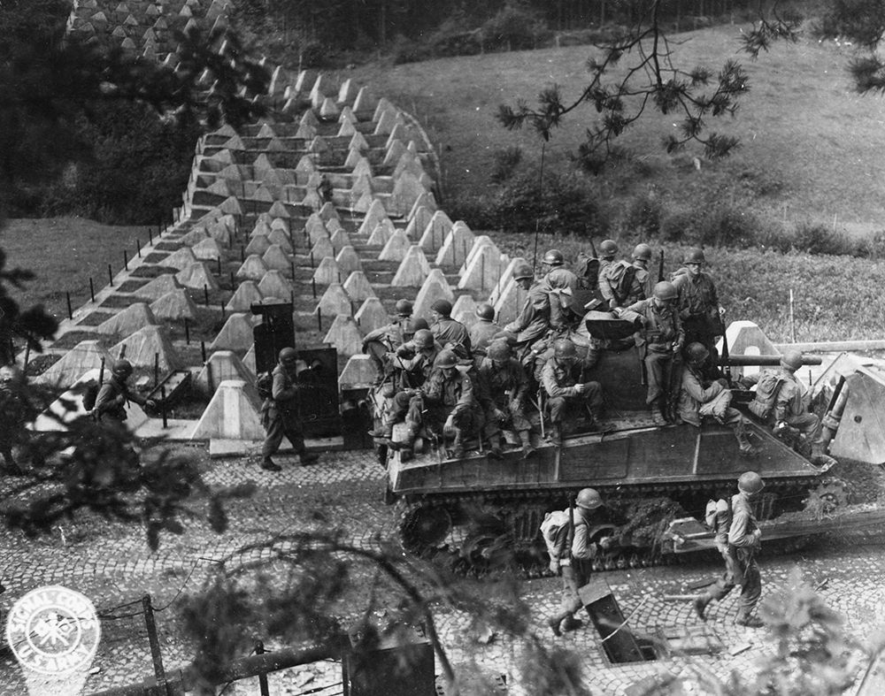 An American tank carrying troops through the German defenses of the Siegfried Line. The pyramidal structures in the background are concrete tank obstructions called “dragon’s teeth.” Pennsylvania State Archives, MG-356
