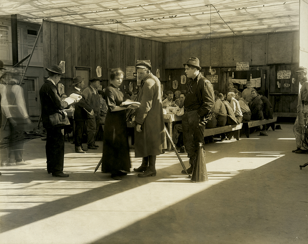On the dining hall set of Sunshine Molly, Weber, with her script in hand, confers with cameraman Dal Clawson while Smalley, dressed as Bull Forrest, stands by with his hand in his pocket. In front of him is the megaphone likely used when directing. Note the fabric “ceiling” at the top of the set to diffuse the California sun. From the Core Collection Production and Biography Files of the Margaret Herrick Library, Academy of Motion Picture Arts and Sciences