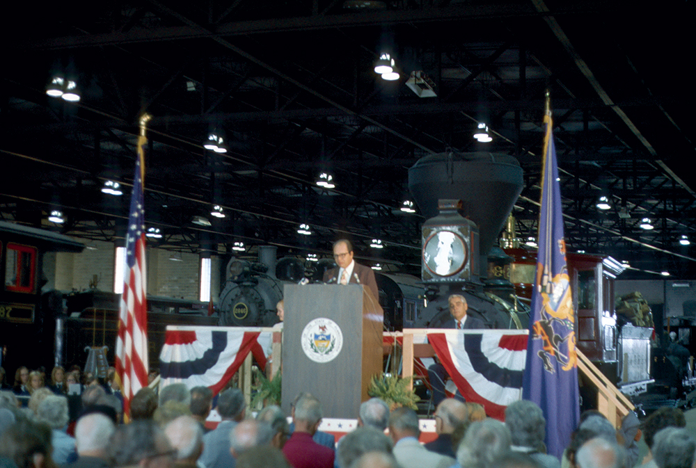 PHMC Executive Director William Wewer speaks before a crowd of visitors gathered in front of the Virginia & Truckee No. 20 Tahoe steam locomotive during the dedication and grand opening ceremony for the Railroad Museum, held on September 10, 1975, with Gov. Milton Shapp in attendance. Rather than cutting a ribbon, the governor rang the polished brass bell of the Tahoe. Railroad Museum of Pennsylvania, PHMC